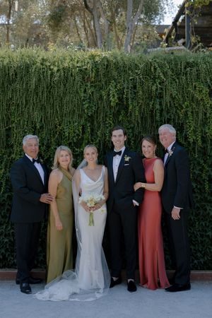 Formal wedding party group photo taken against a lush green hedge backdrop.