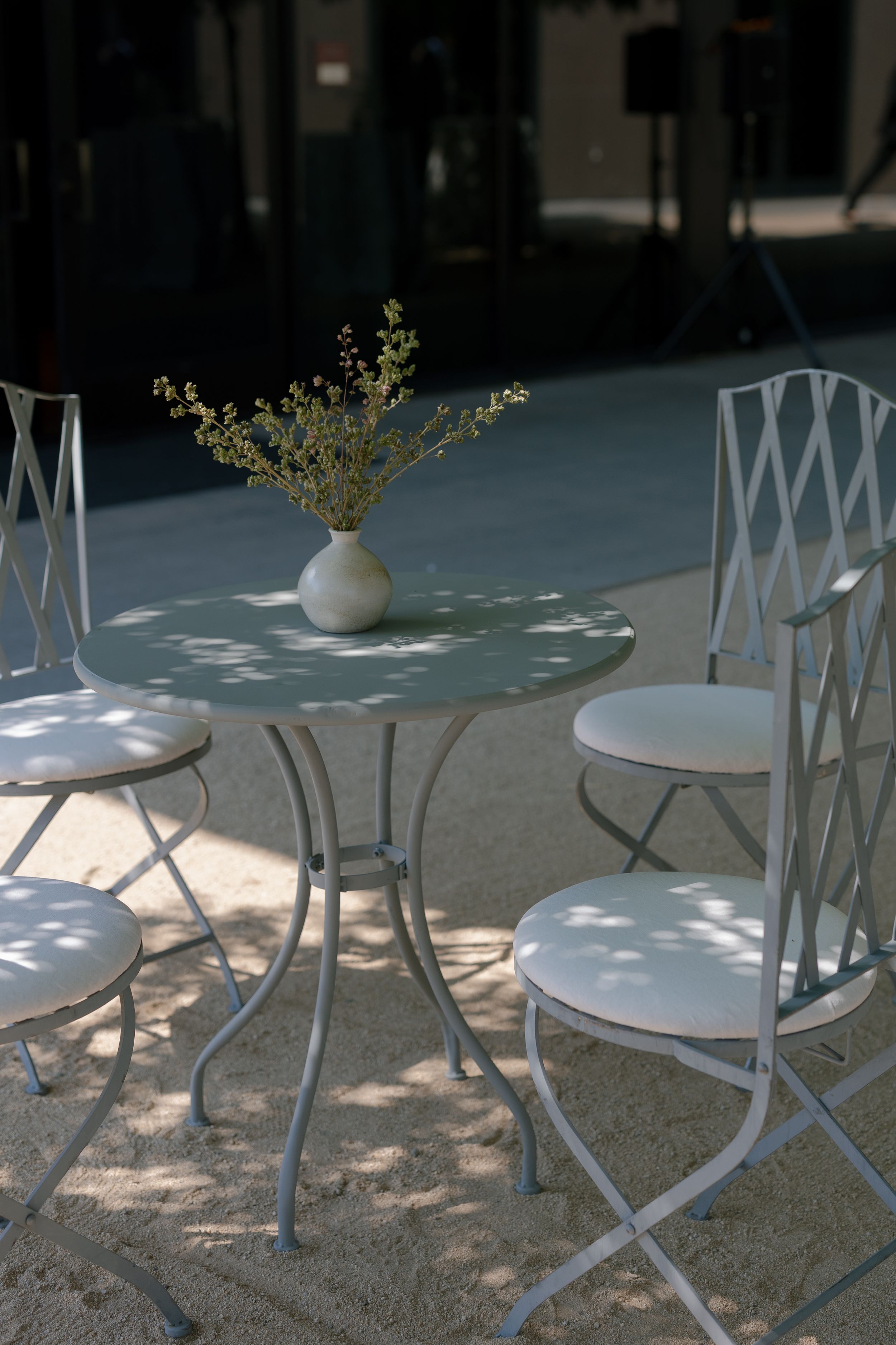 White metal cafe tables and chairs arranged on a patio with dappled sunlight filtering through trees.