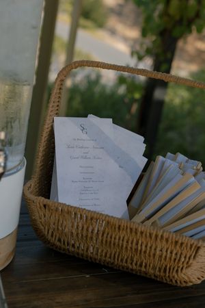 White paper wedding programs displayed in a woven basket on a dark wooden surface with blurred background.