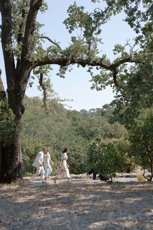 Wedding guests walk along a scenic hillside path under large oak trees on a sunny afternoon.