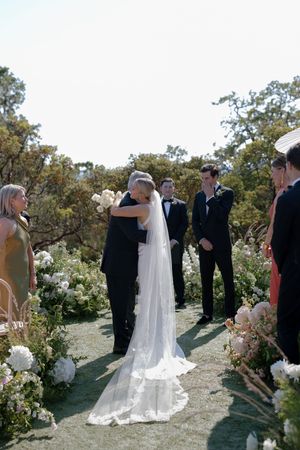 A wedding ceremony takes place on a hilltop surrounded by lush greenery and floral arrangements.
