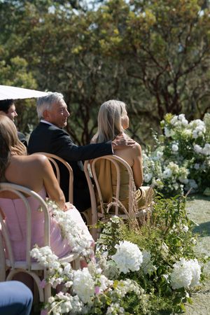 Wedding guests seated in wooden chairs surrounded by white floral arrangements during an outdoor garden ceremony.