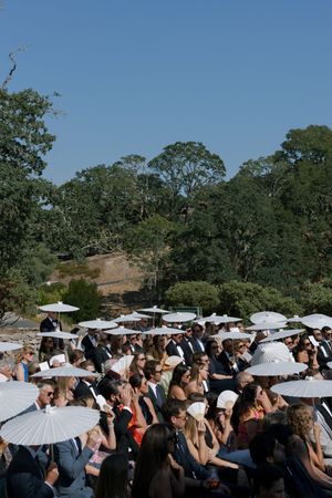 Wedding guests with white parasols seated outdoors under clear blue skies with trees in background.