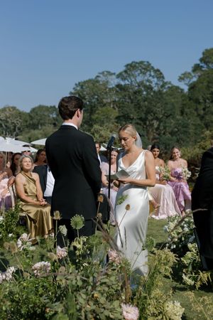 Wedding guests gather outdoors in a garden setting with lush greenery and trees in the background.