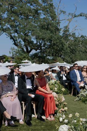 Wedding guests with parasols gather outdoors under oak trees for an elegant garden ceremony.