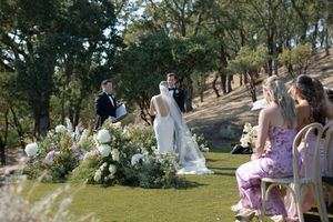 Outdoor wedding ceremony setup with white chairs and floral arrangements in a wooded natural setting.