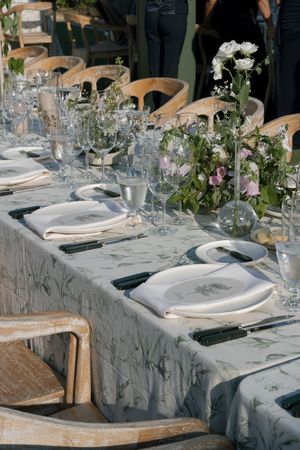 Elegant table setting with white floral centerpieces, wooden chairs, and crystal glassware on a light gray tablecloth.