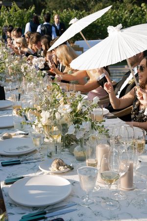 Long white dining table set outdoors with paper parasols, delicate floral arrangements and place settings.