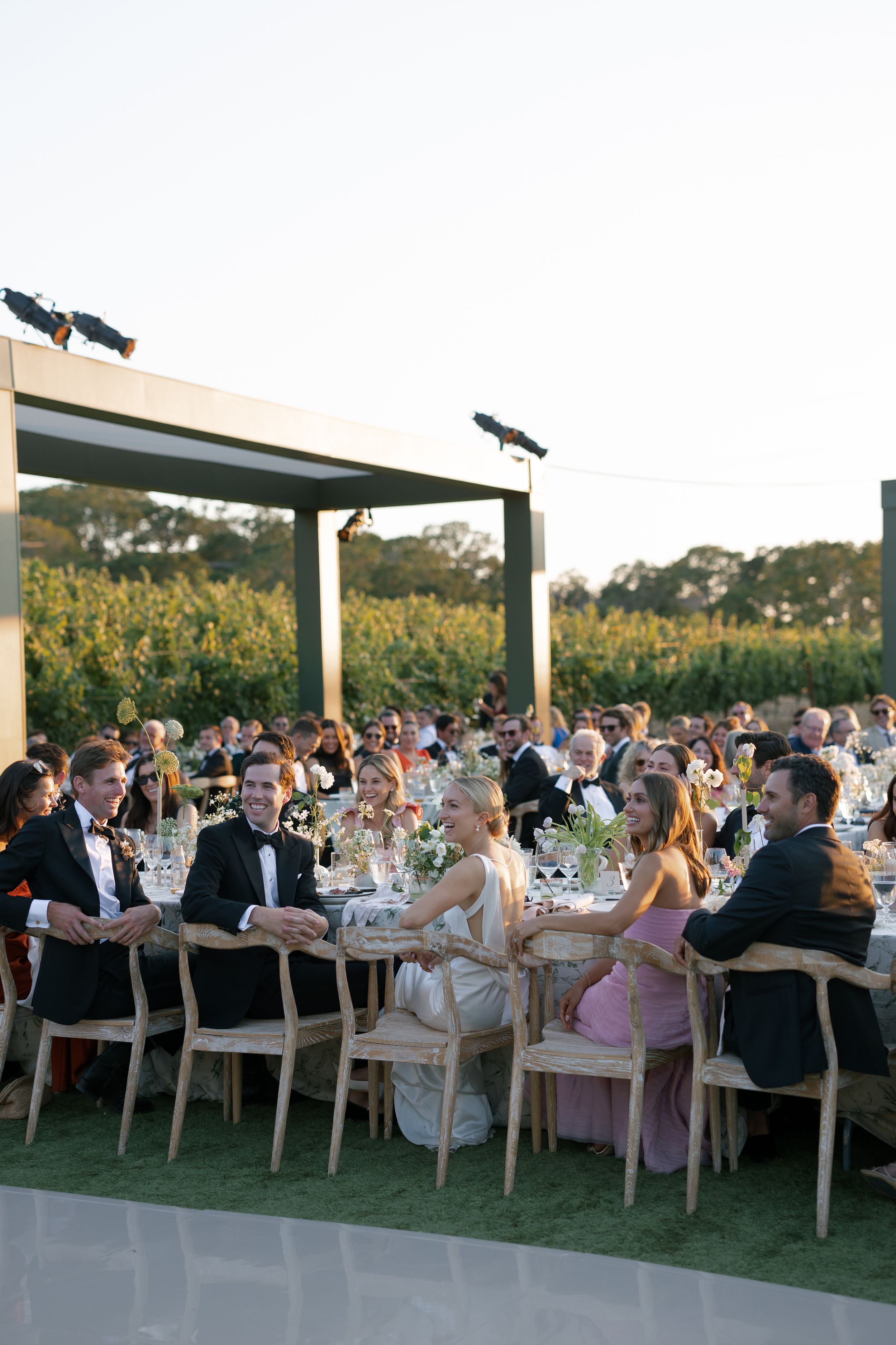 Outdoor wedding reception with guests seated at long tables under wooden pergolas at sunset.