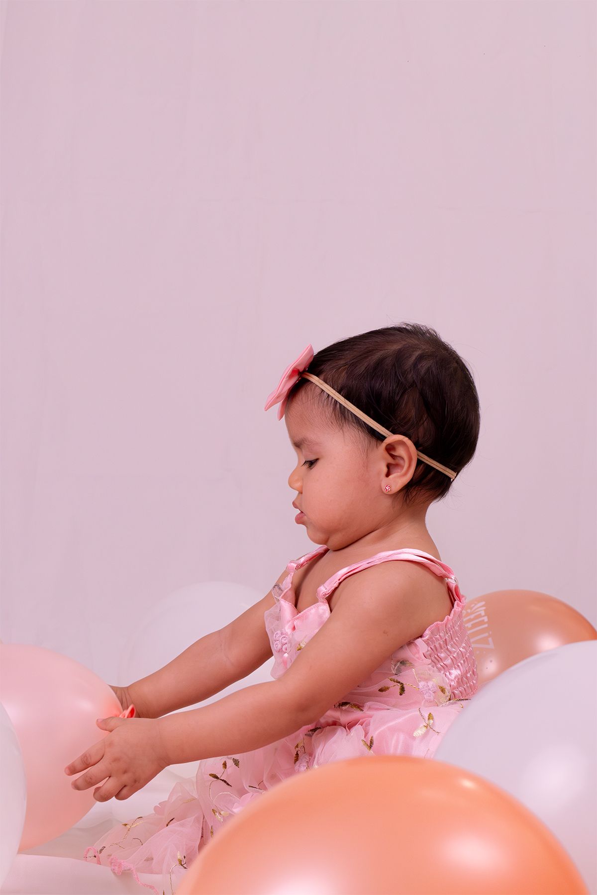 Baby girl in pink floral dress surrounded by balloons, viewed from behind with hair accessory.