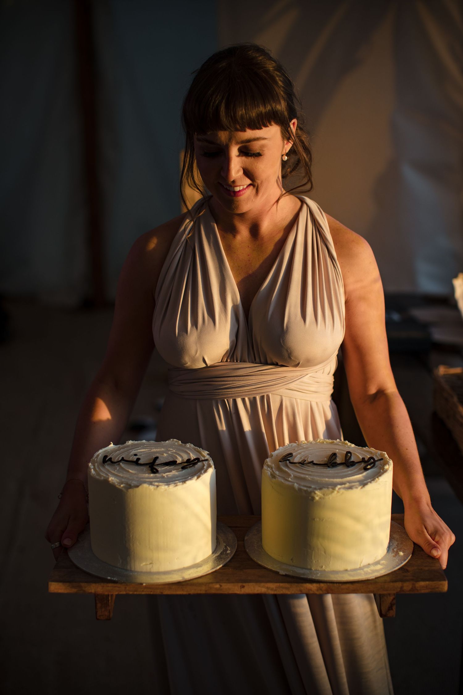 Smiling woman in elegant halter dress holding wooden board with two white frosted cakes at a celebration.