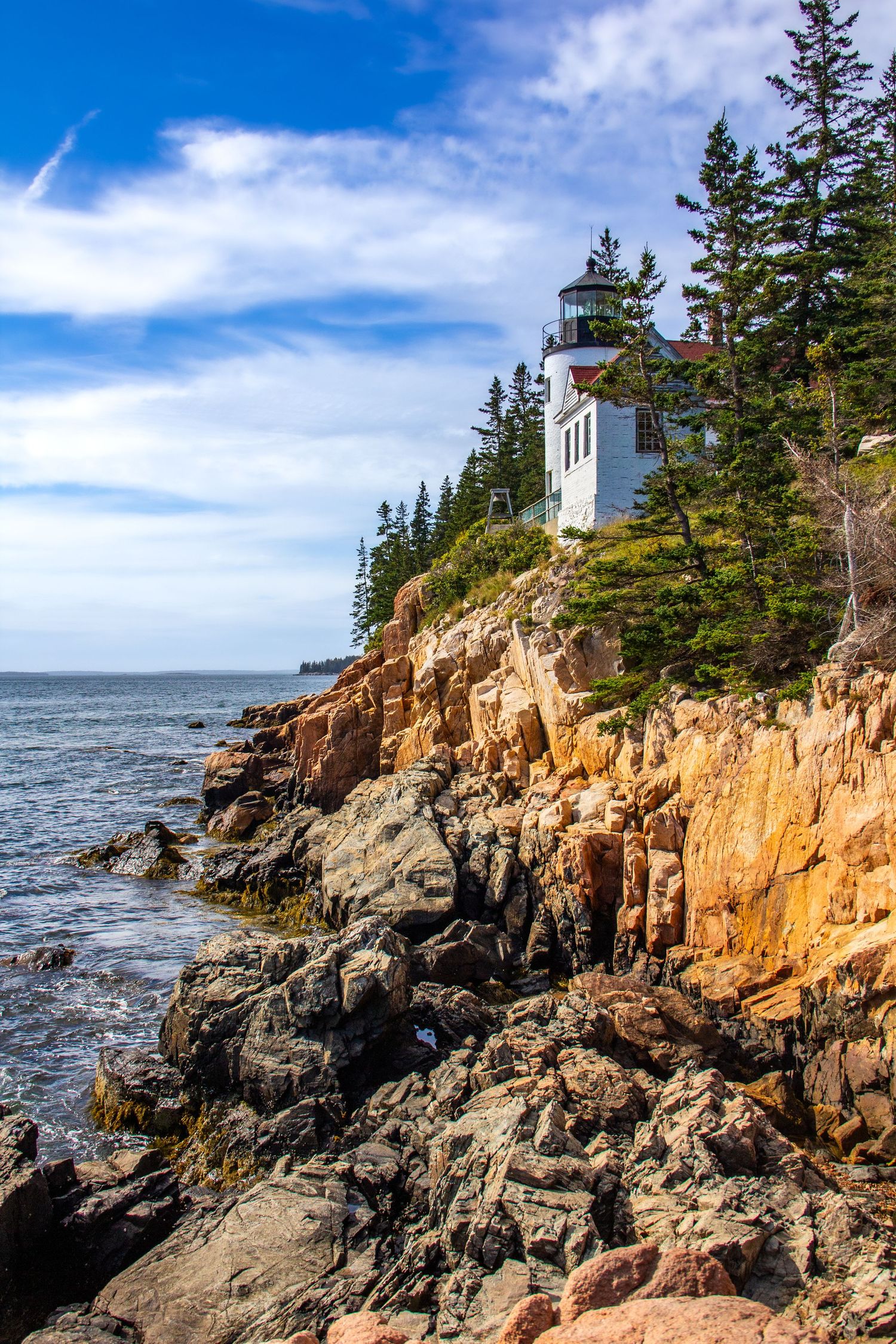Bass Harbor Head Lighthouse perched on rugged red rocky cliffs surrounded by evergreen trees under a vivid blue sky in Maine.