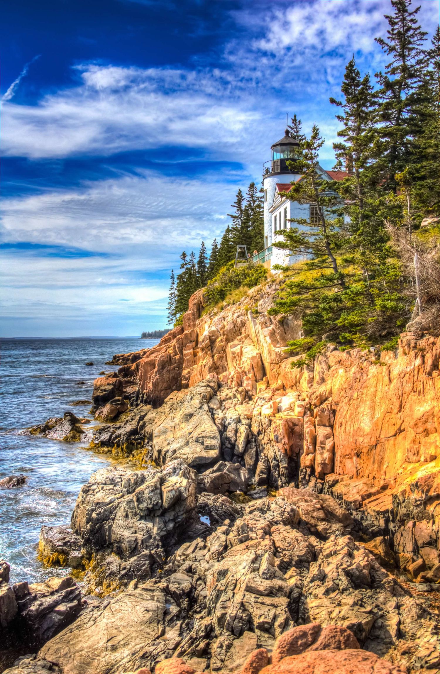 Bass Harbor Head Lighthouse perched on rugged red rocky cliffs surrounded by evergreen trees under a vivid blue sky in Maine.