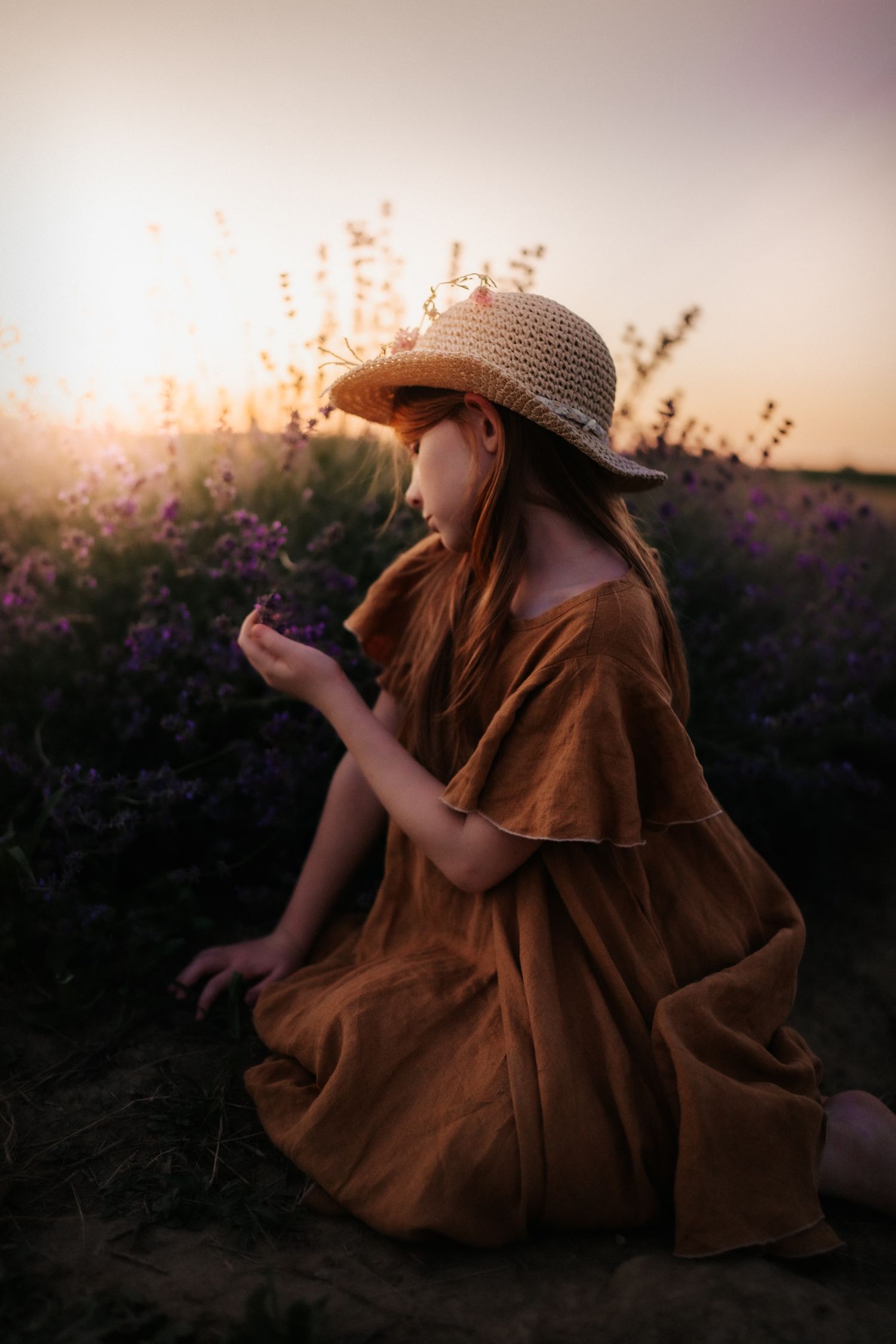 A young girl in a brown dress and straw hat sits in a lavender field at golden hour, smelling flowers.