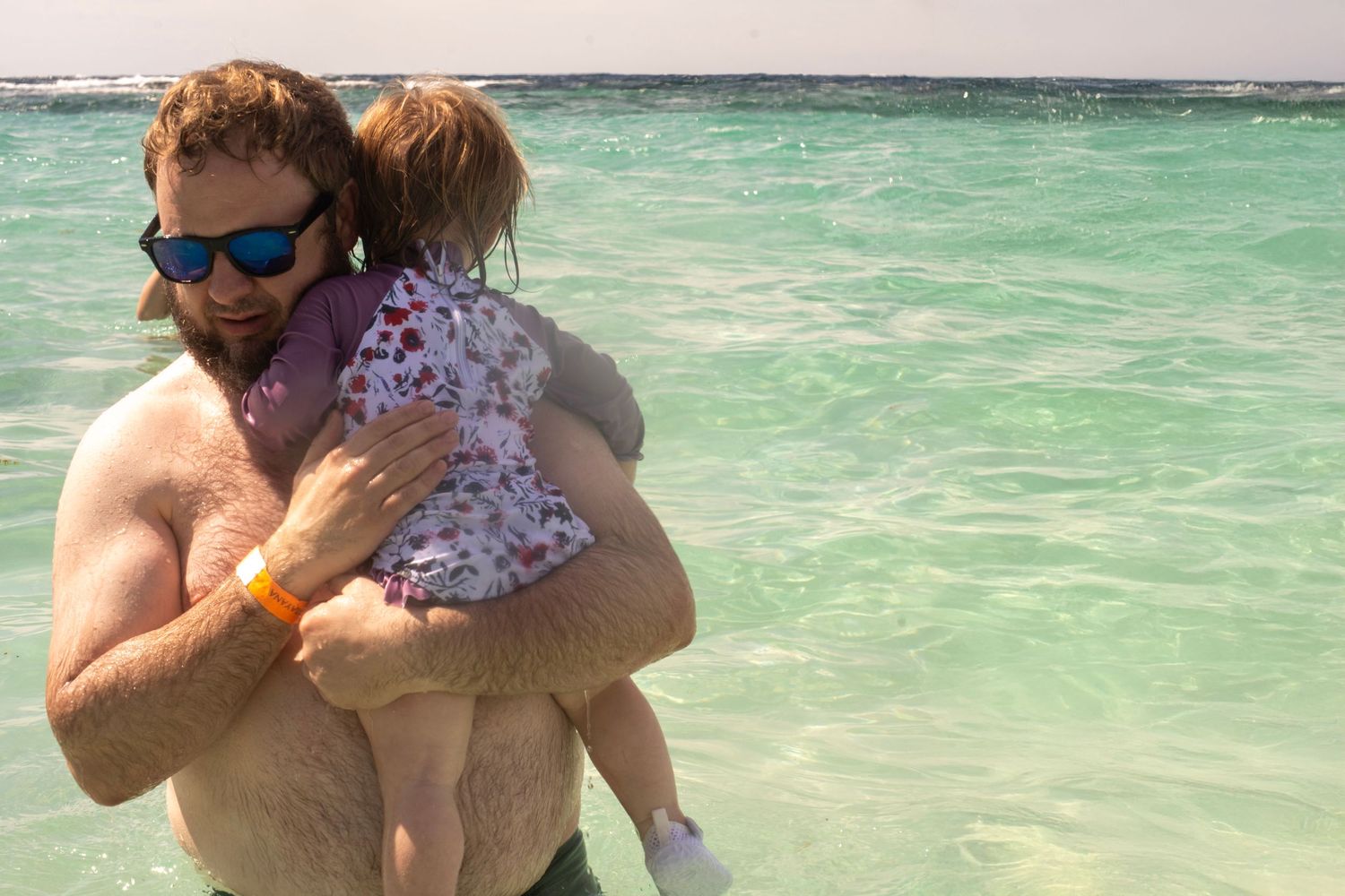A shirtless man wearing sunglasses holds a young child at a tropical beach with clear turquoise water.