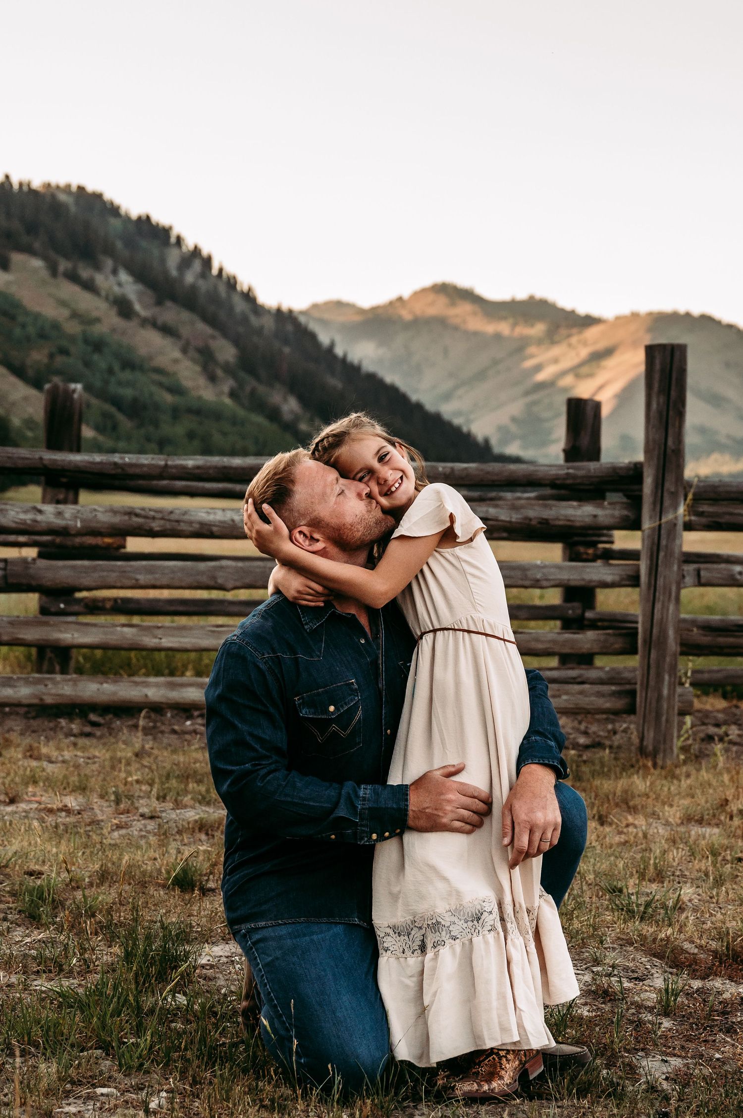 A father kneels in a mountain meadow, kissing his smiling daughter in a white dress near a rustic wooden fence.