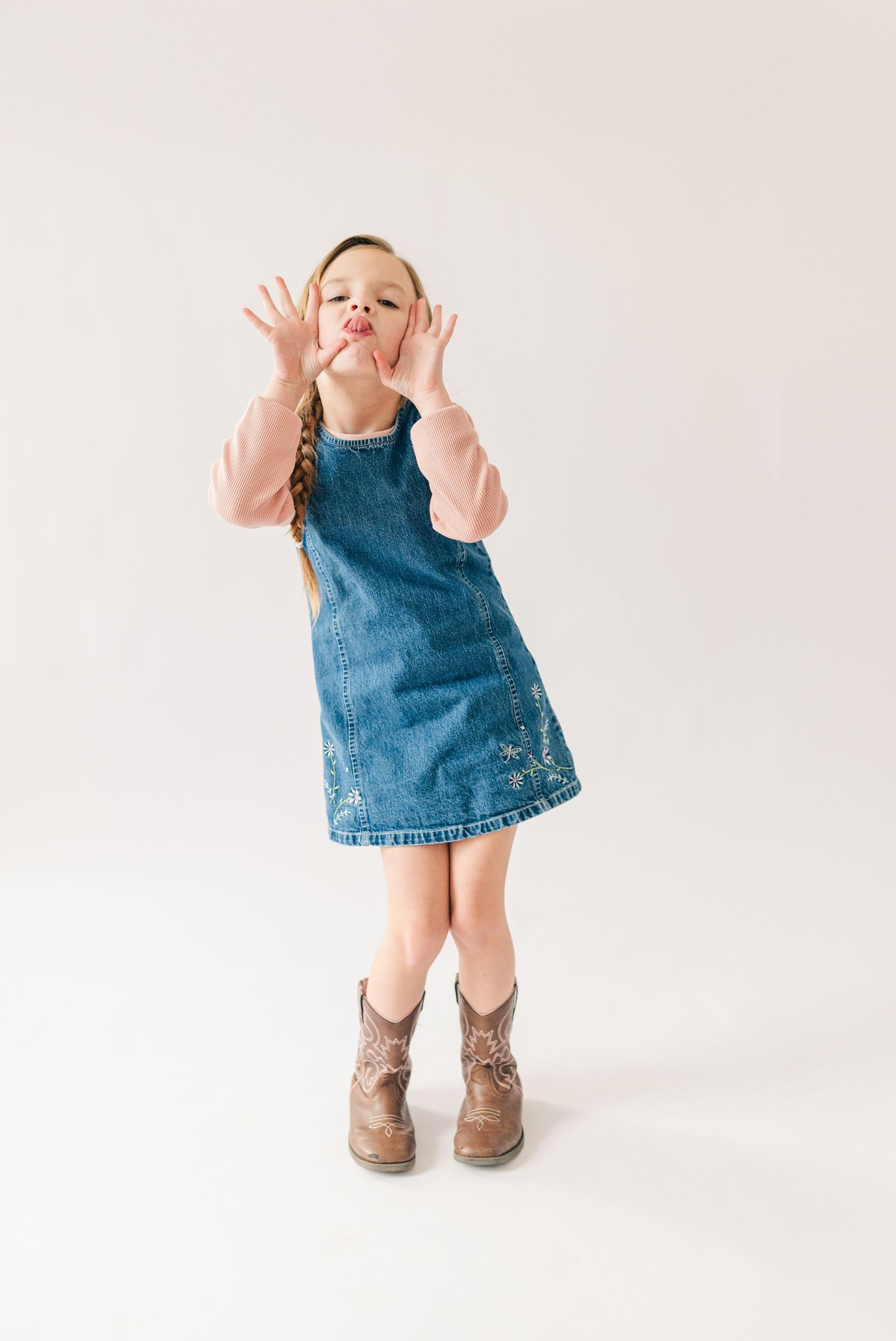 Small child in denim dress and boots making playful gestures against white background.