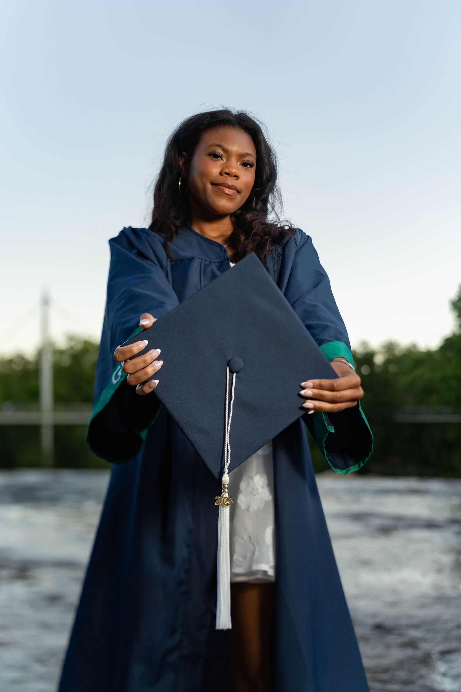 Graduate in navy blue cap and gown holds diploma while posing outdoors by water.