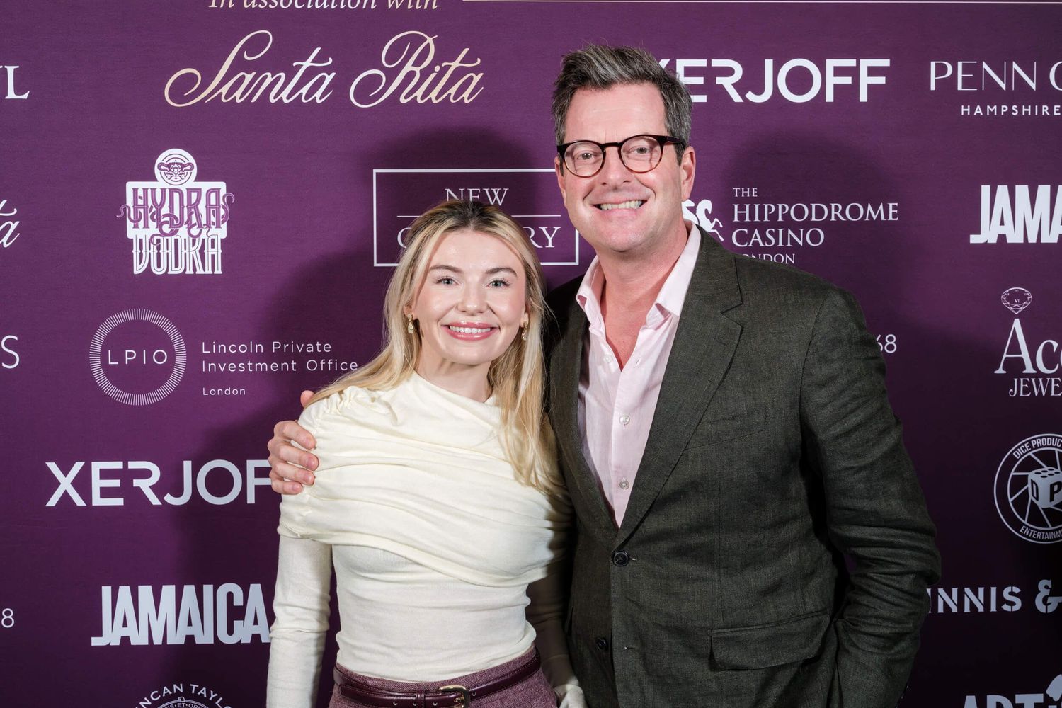 Two people pose together at a formal event with purple backdrop and sponsor logos visible behind them.