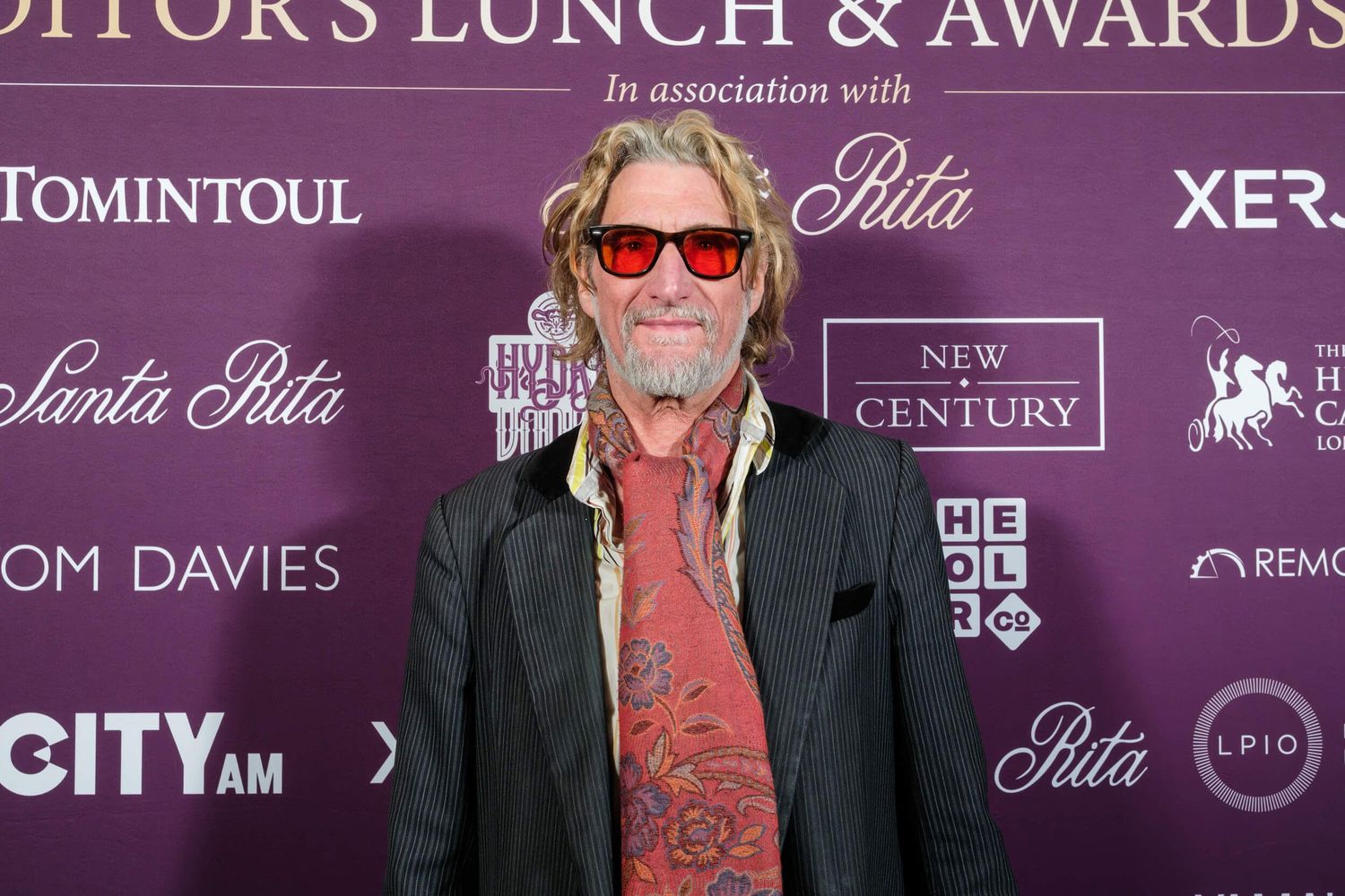 Person wearing sunglasses and dark jacket poses on red carpet at awards event.