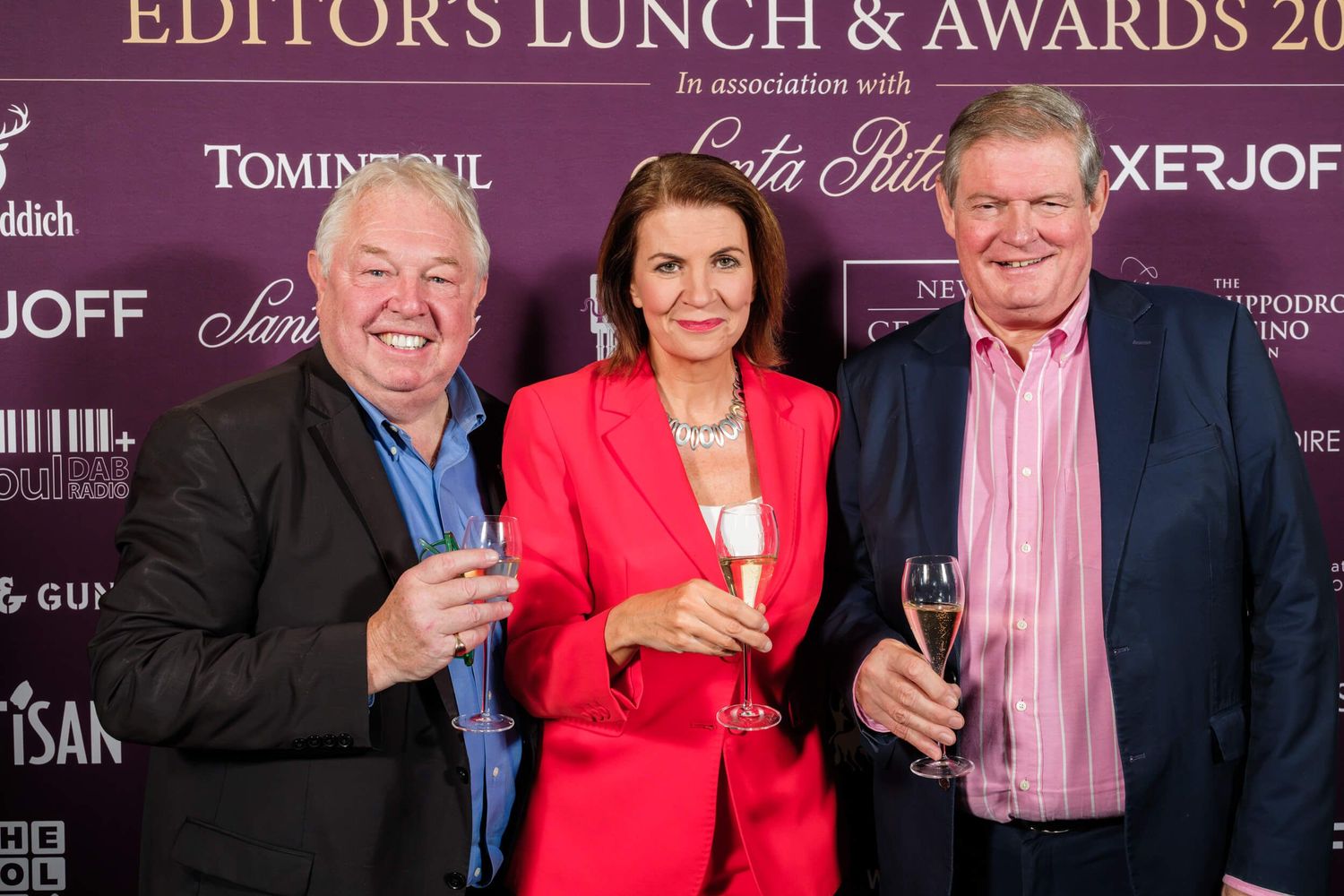 Three guests in formal attire smile while holding drinks at an awards ceremony.