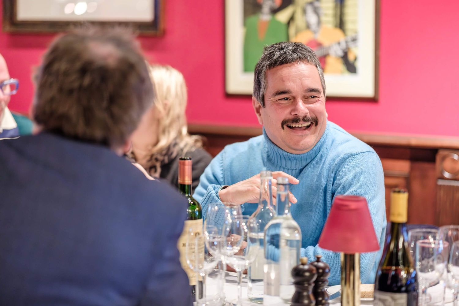 A group of people enjoying drinks and conversation at a restaurant with red walls and framed artwork.