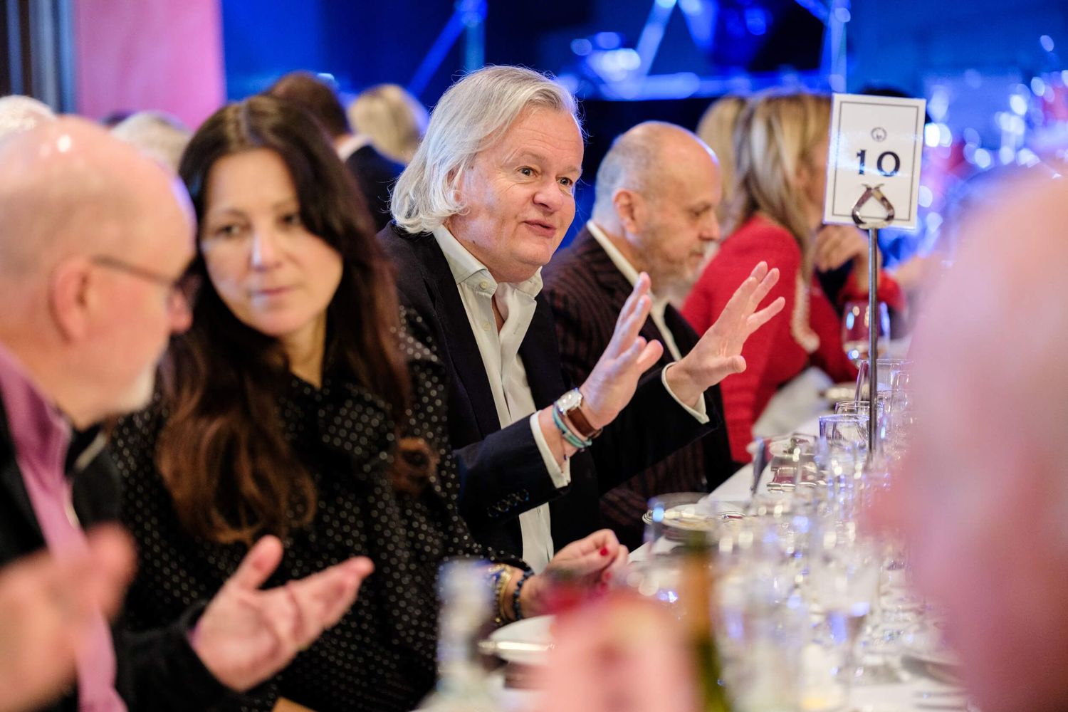 People seated at an elegant dinner table with blue lighting applaud during an upscale event.