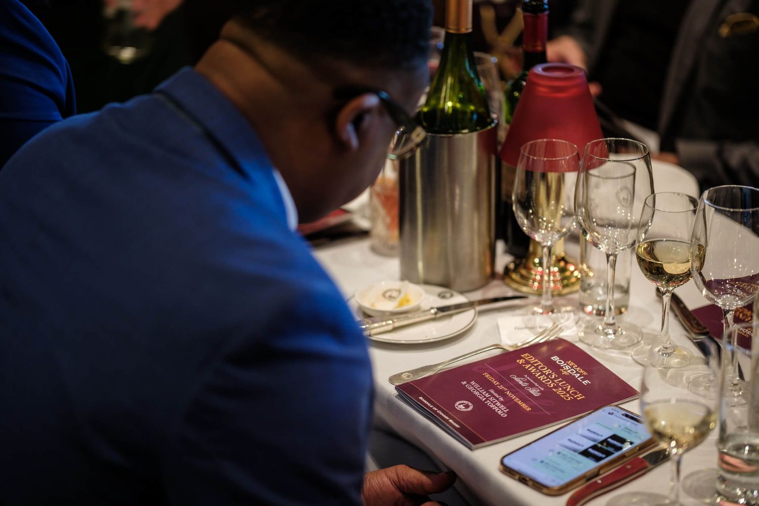 Someone in a blue jacket reviews a menu at an elegant dinner table set with wine glasses and bottles.