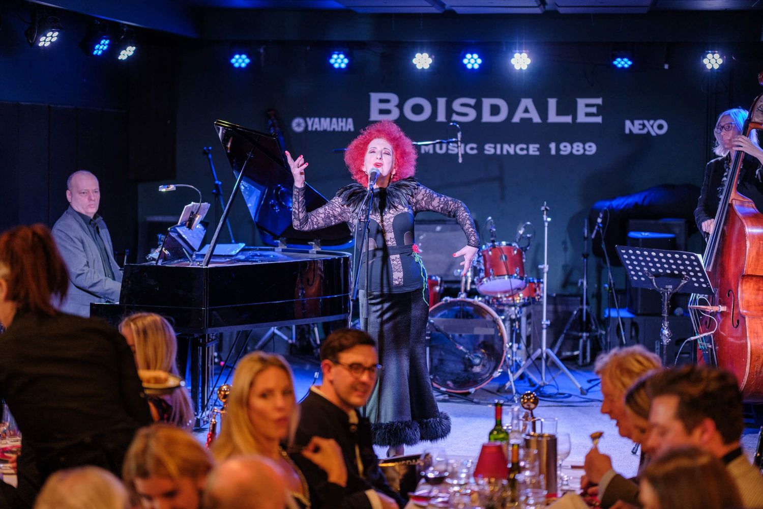 Musicians perform on stage at a live music venue with blue lighting and instruments visible in the background.