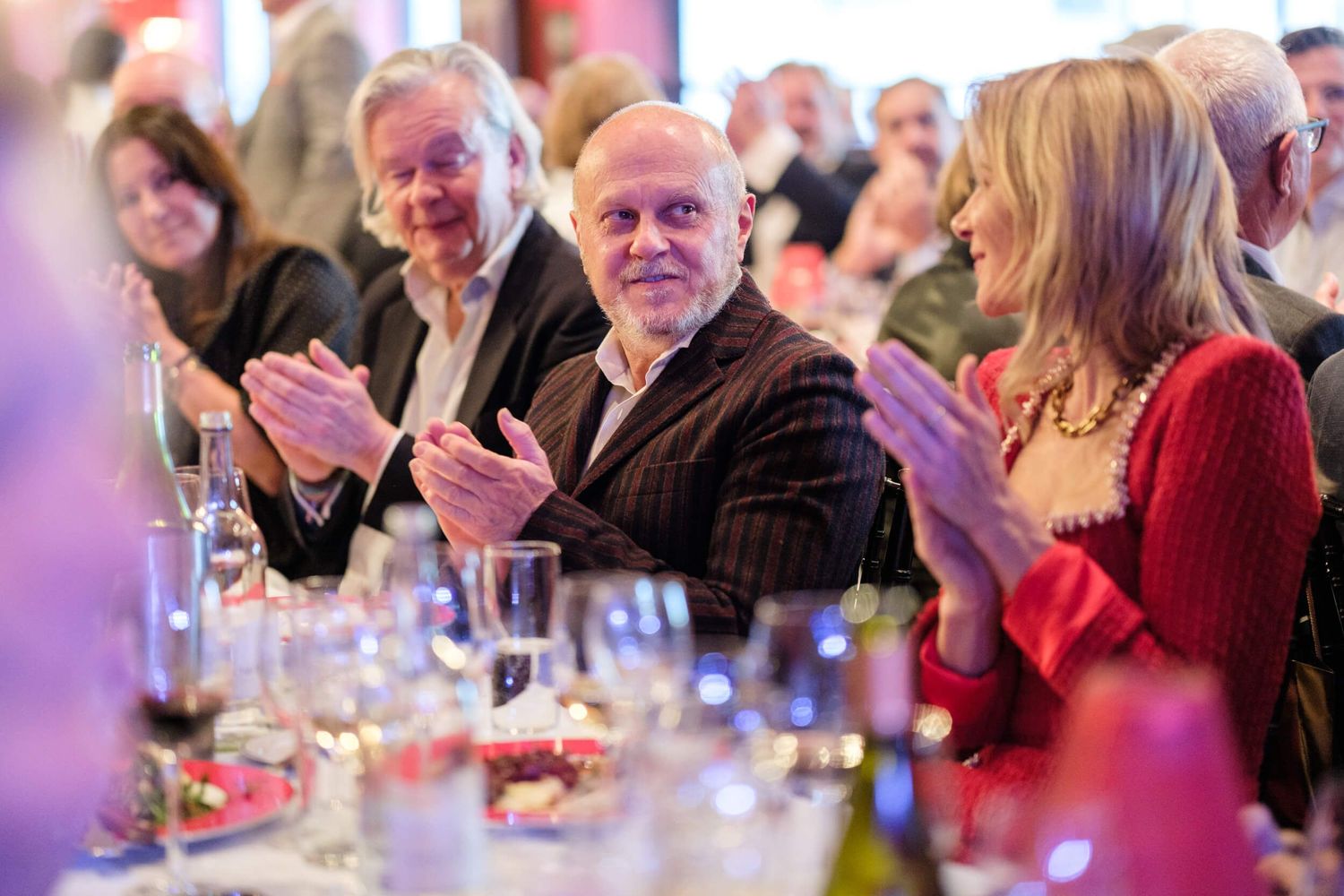 People applaud during an elegant dinner event with wine glasses visible on tables in a warmly lit room.