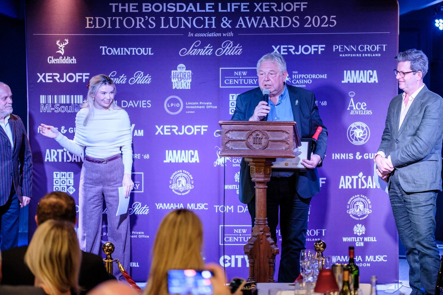 People seated at tables during an awards ceremony with purple lighting and branded backdrop displays.