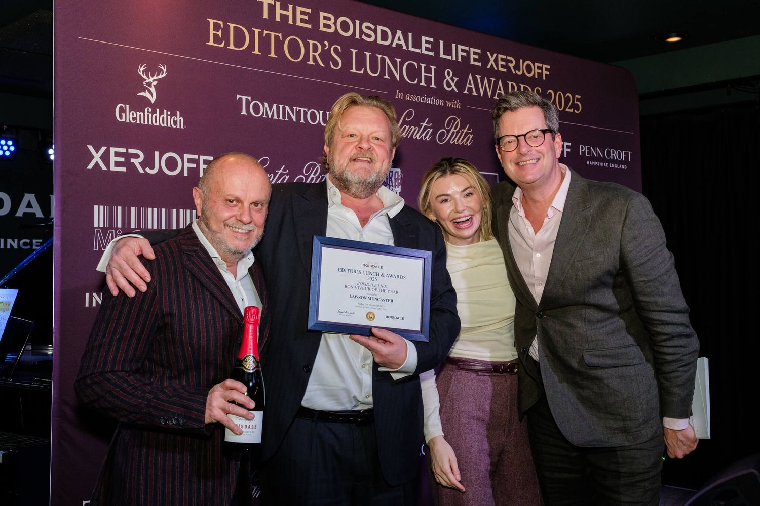 Award winners celebrate with drinks and certificates at an evening ceremony against a purple-lit backdrop.