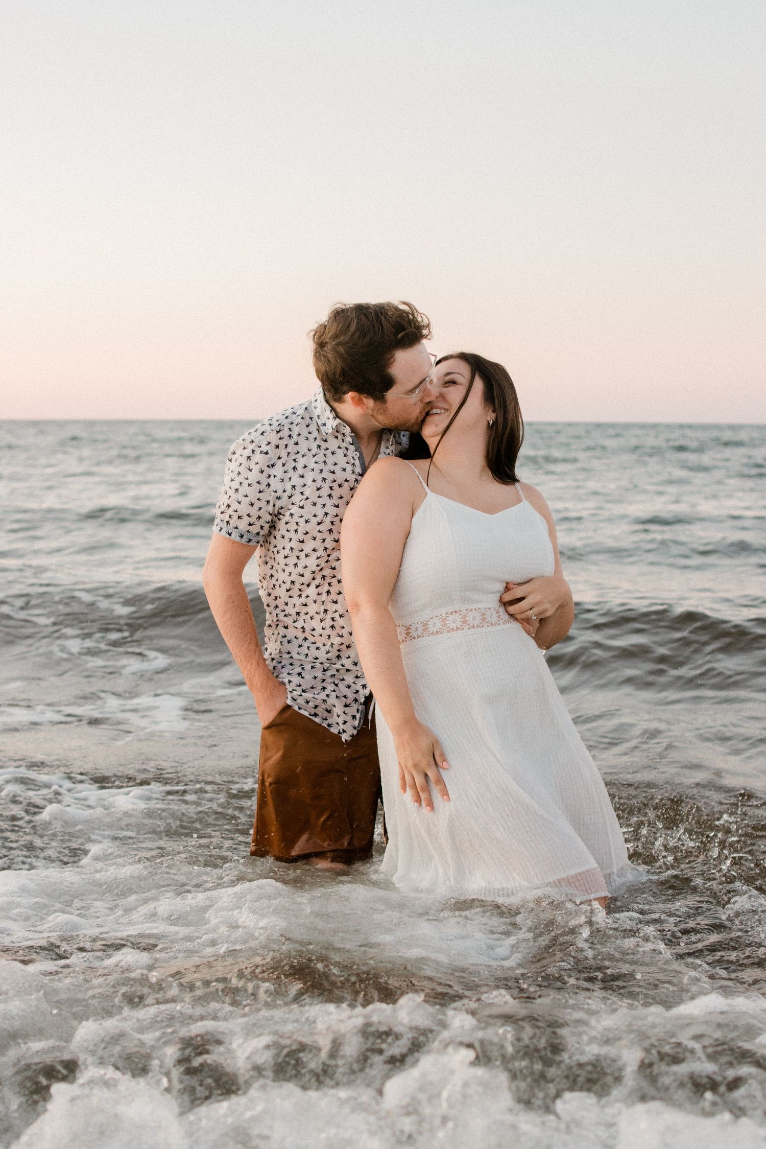Romantic couple embraces in ocean waves at sunset, with person wearing floral shirt and white dress standing in shallow water.