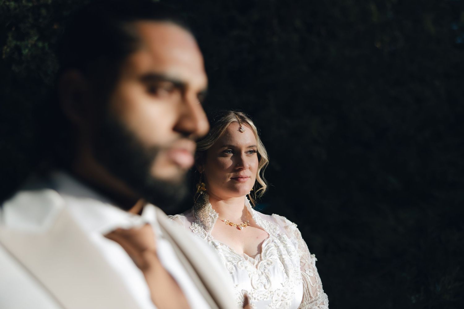 Artistic wedding portrait of a couple in white attire against a dark background with dramatic lighting.