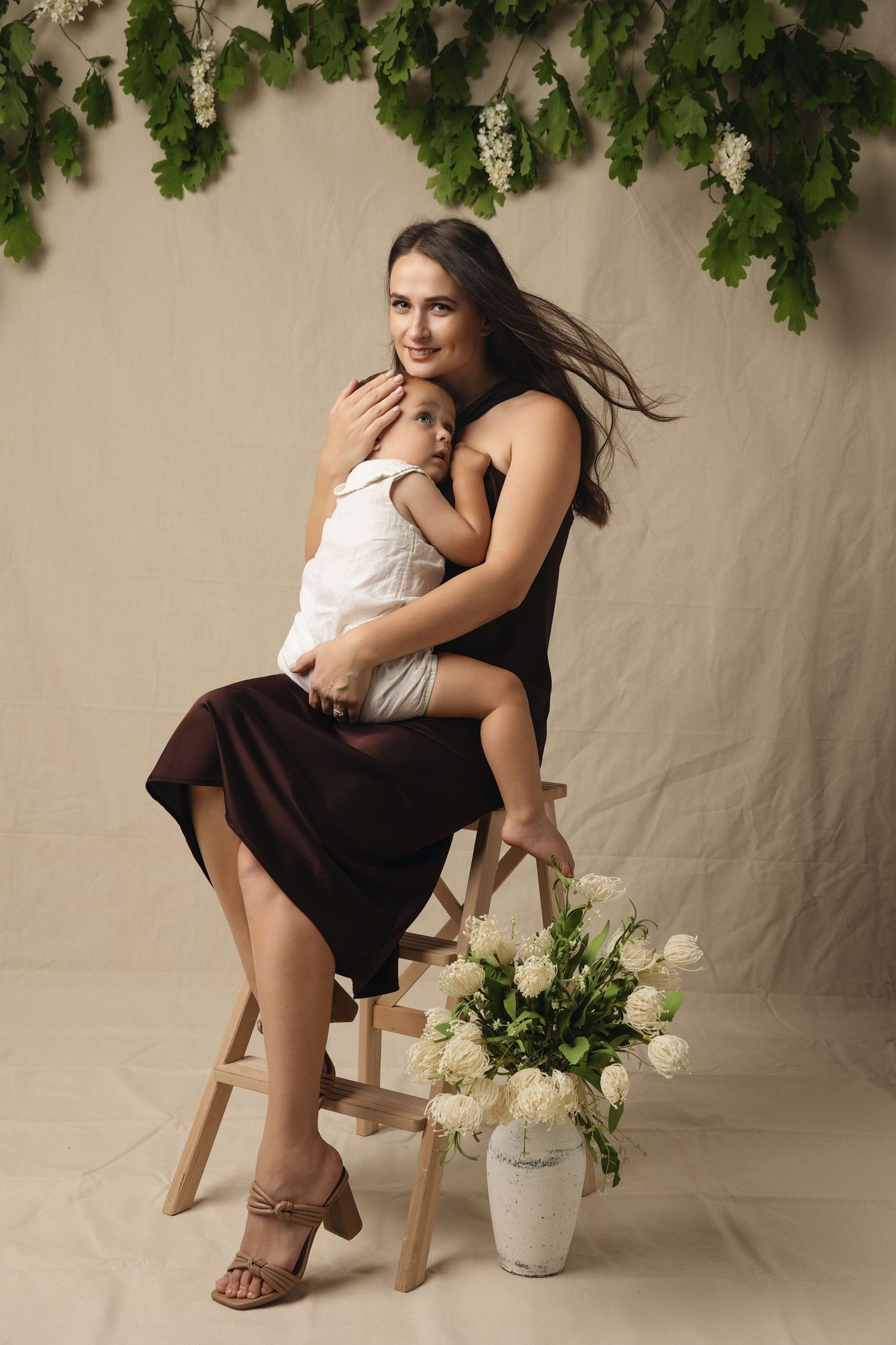 A tender studio portrait of a parent and baby in black outfits against a brown backdrop, seated on a white stool.