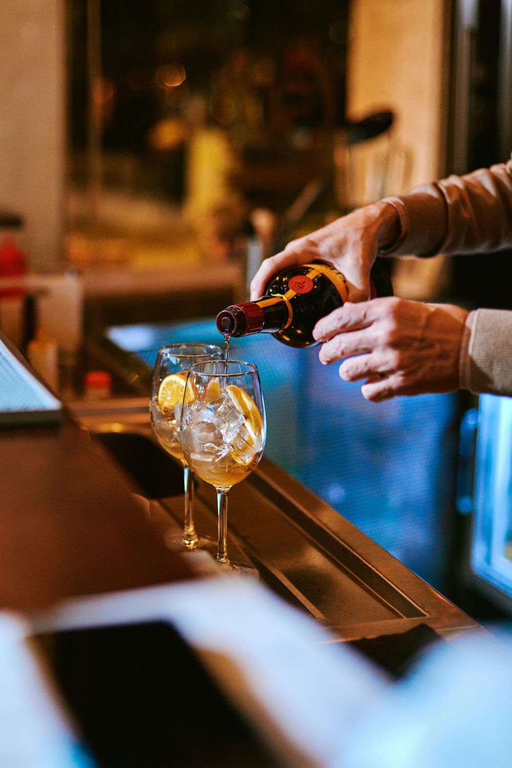 Bartender pouring beer from bottle into wine glass with ice and lemon slices at bar.