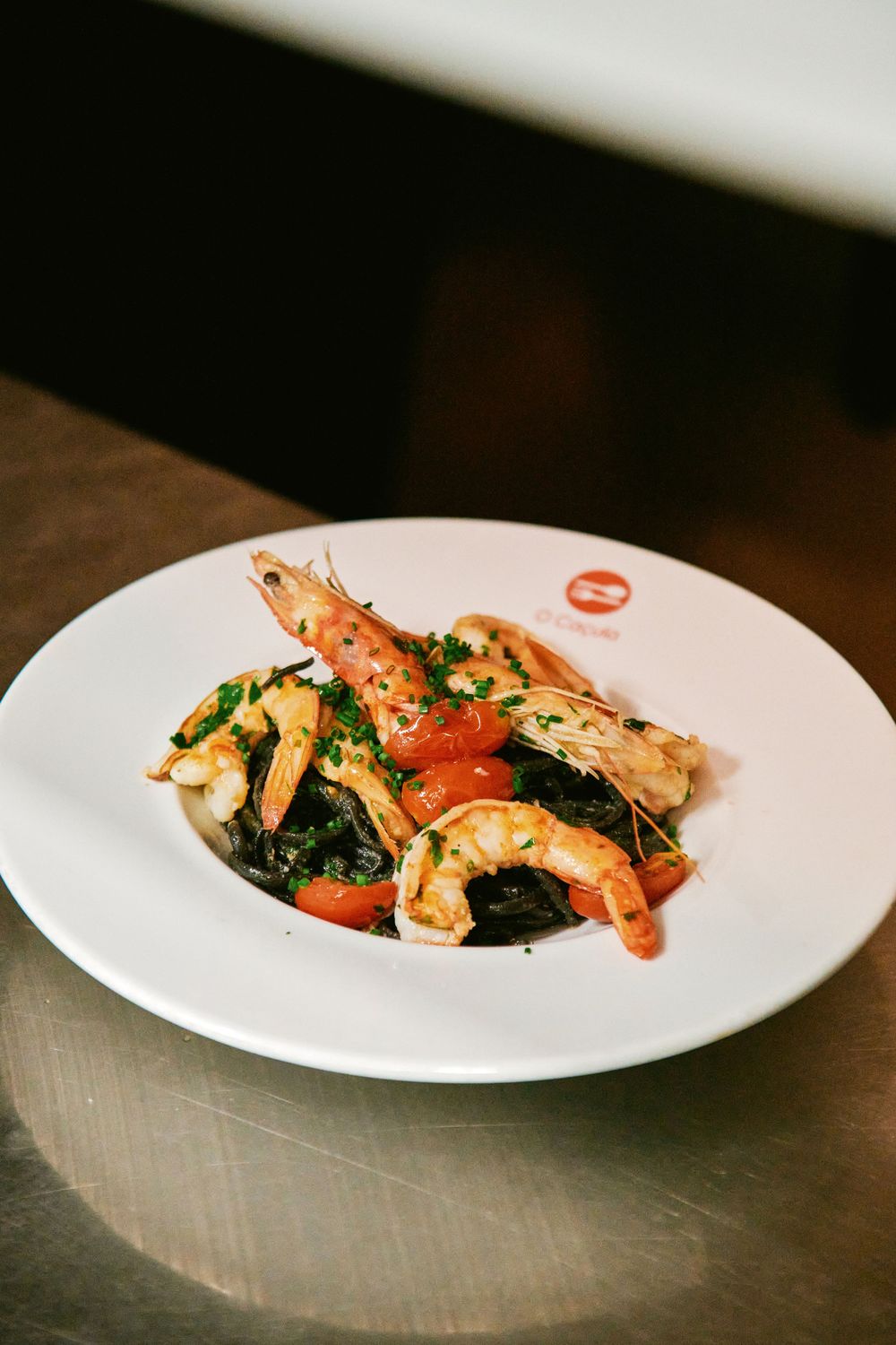 Elegant shrimp dish with black pasta, tomatoes, and herbs served on white plate in restaurant.