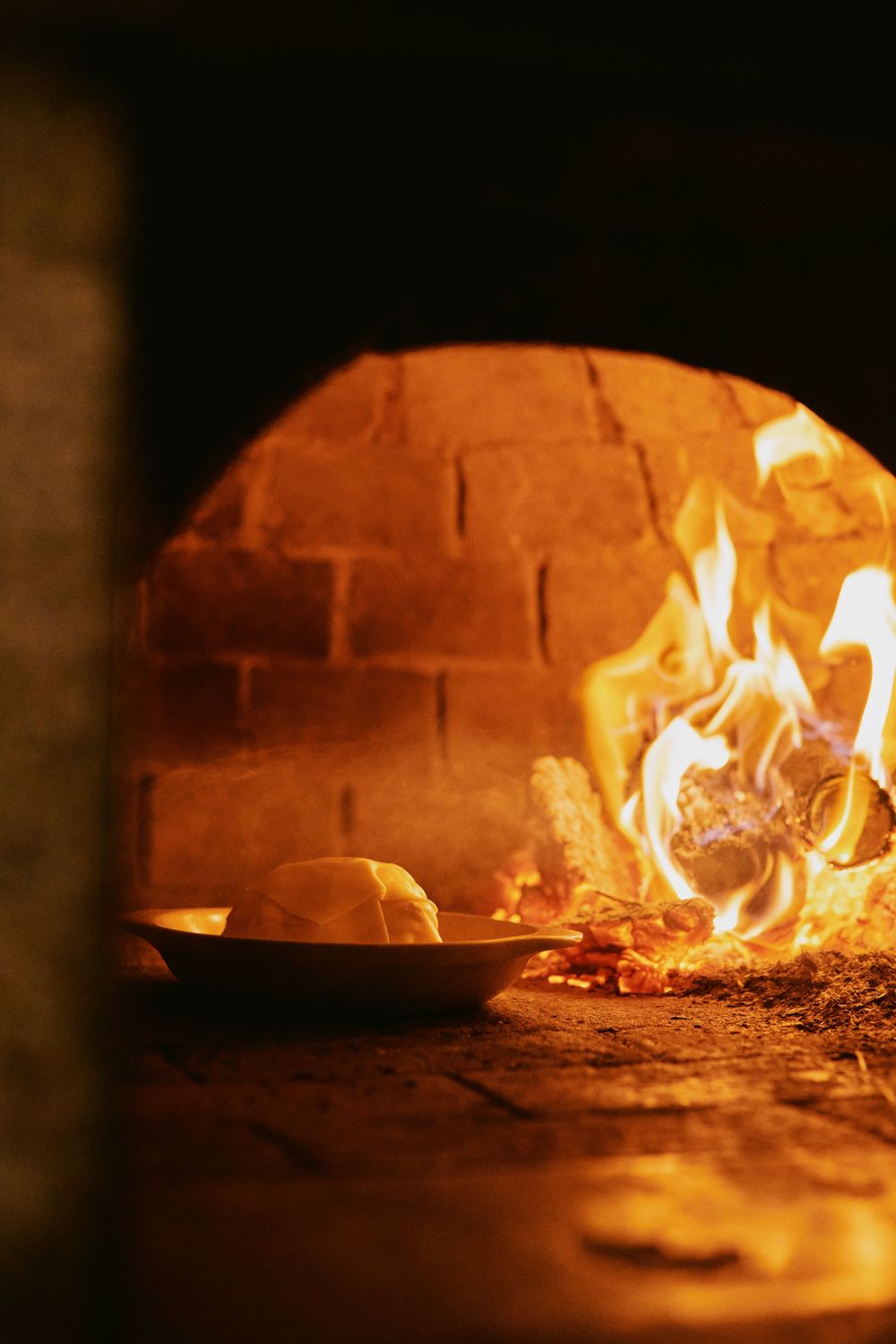 Fresh dough in white dish placed at entrance of glowing wood-fired brick oven with flames.