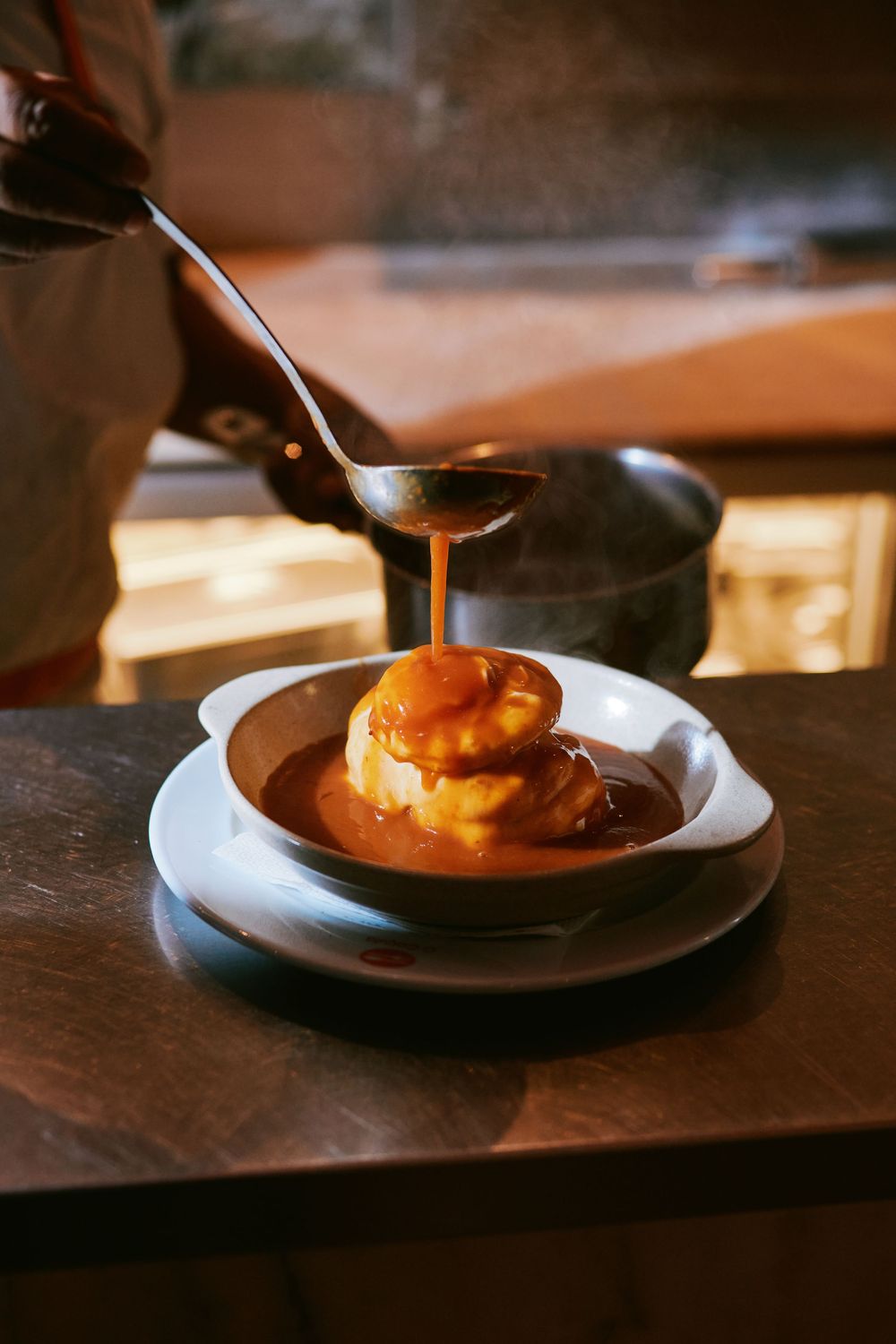 Chef pouring rich brown gravy sauce over a plated dish in a warm restaurant kitchen.