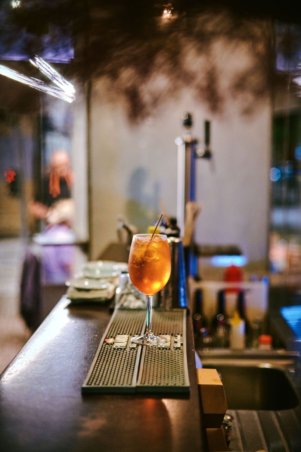 Orange iced cocktail with straw sitting on bar counter in dimly lit restaurant setting.
