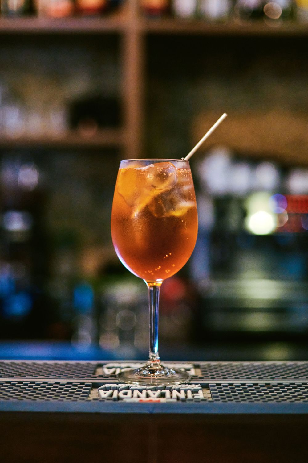 Amber-colored iced cocktail in wine glass with straw on Finlandia bar mat at a bar.