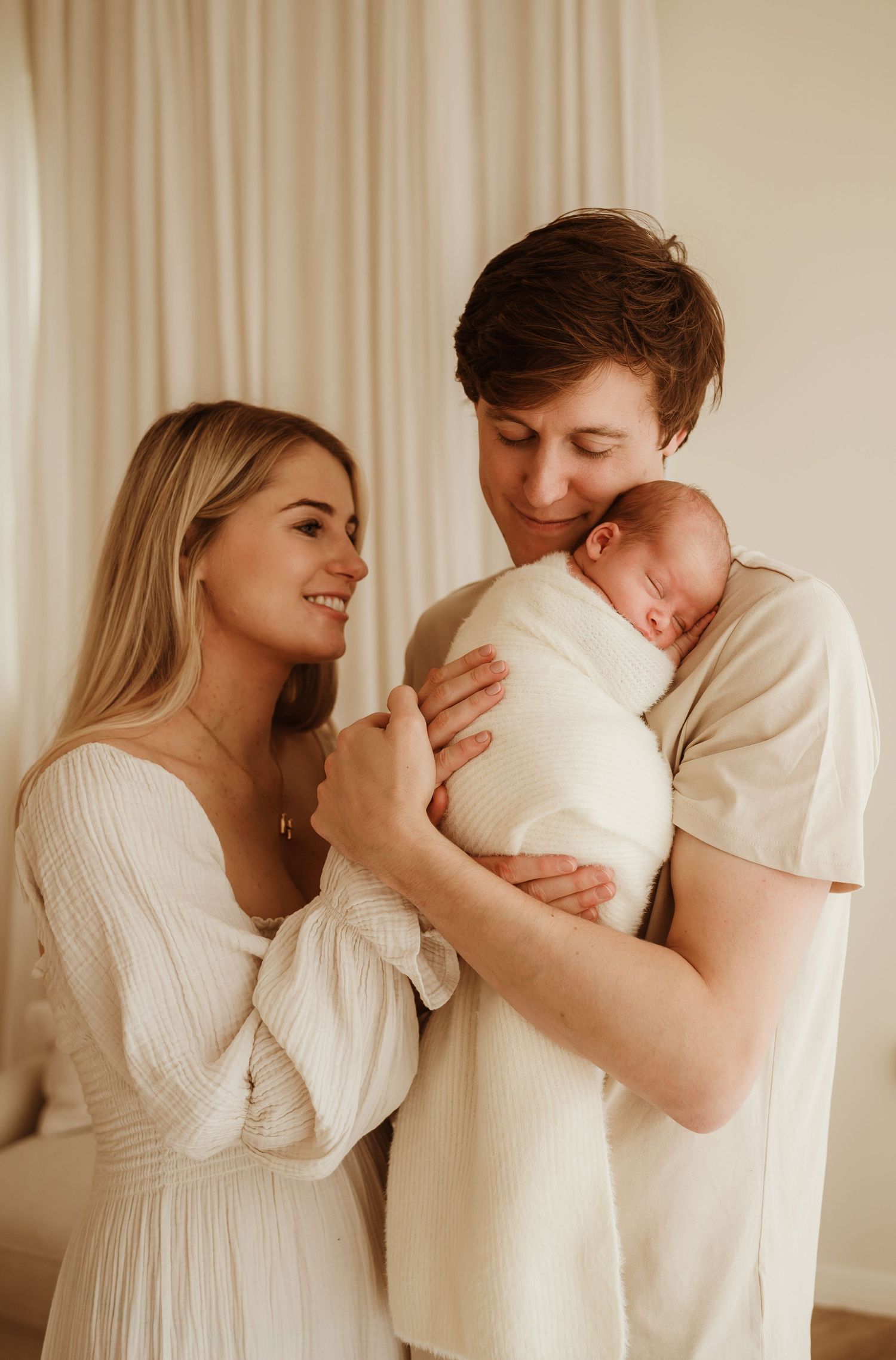A tender family moment as parents share a loving look while holding their newborn baby wrapped in white.