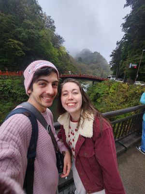 Two people pose together on a foggy day near a red bridge railing in a park setting.