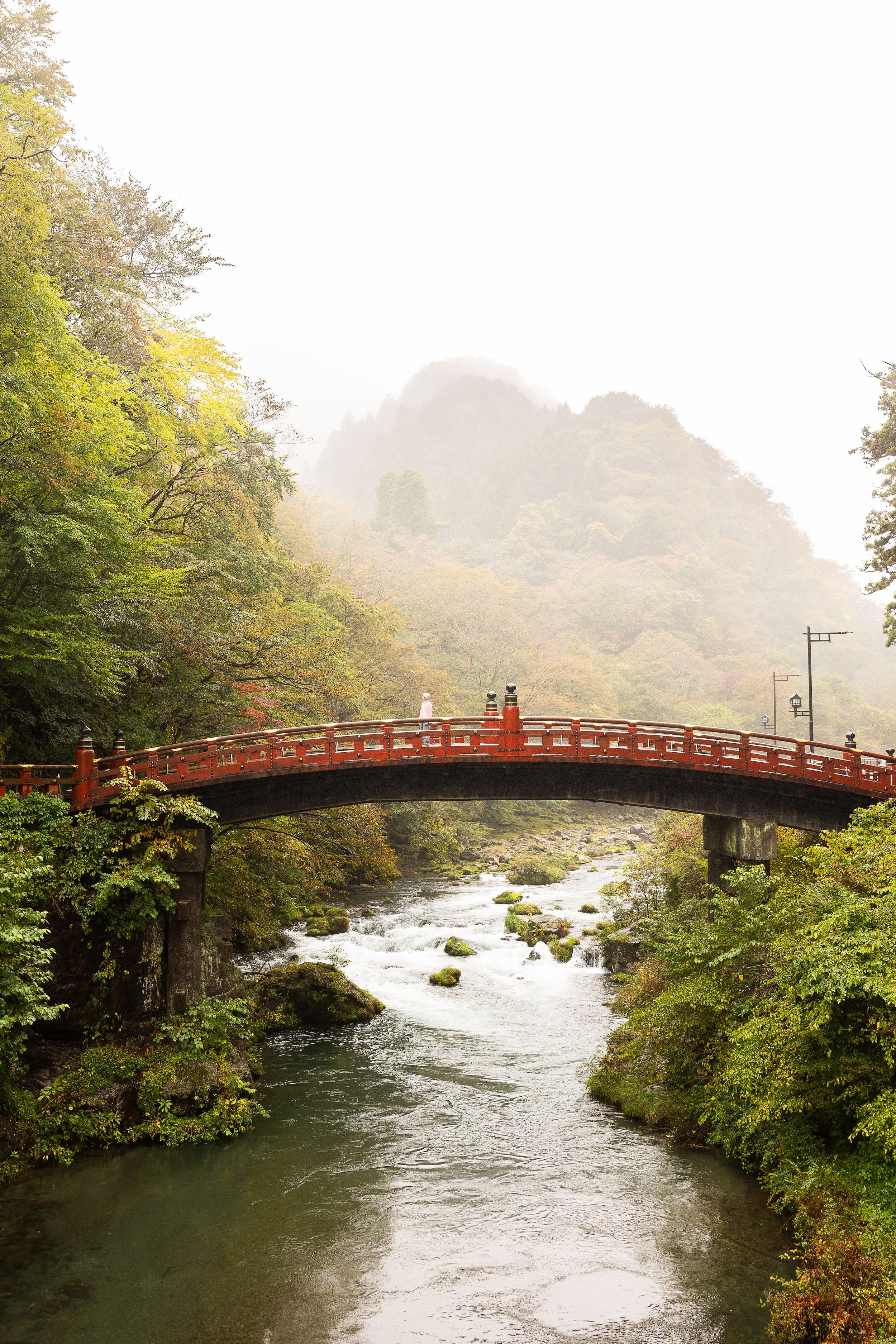 A traditional red Japanese bridge spans a misty river surrounded by lush autumn foliage in Nikko, Japan.