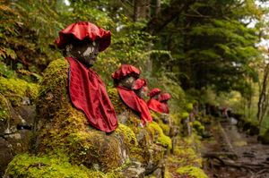 Red-robed statues covered in moss line a stone path through a lush forest in Japan.