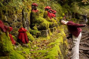 A person in red clothing walks along a mossy forest trail with bright green foliage surrounding them.