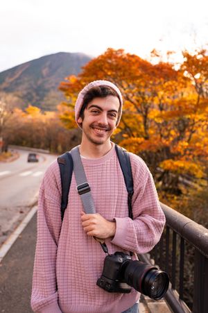 Photographer in pink sweater holding camera poses along autumn road with vibrant orange foliage in background.