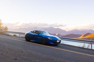 Blue sports car parked on mountain road at sunset with scenic mountain range in background.
