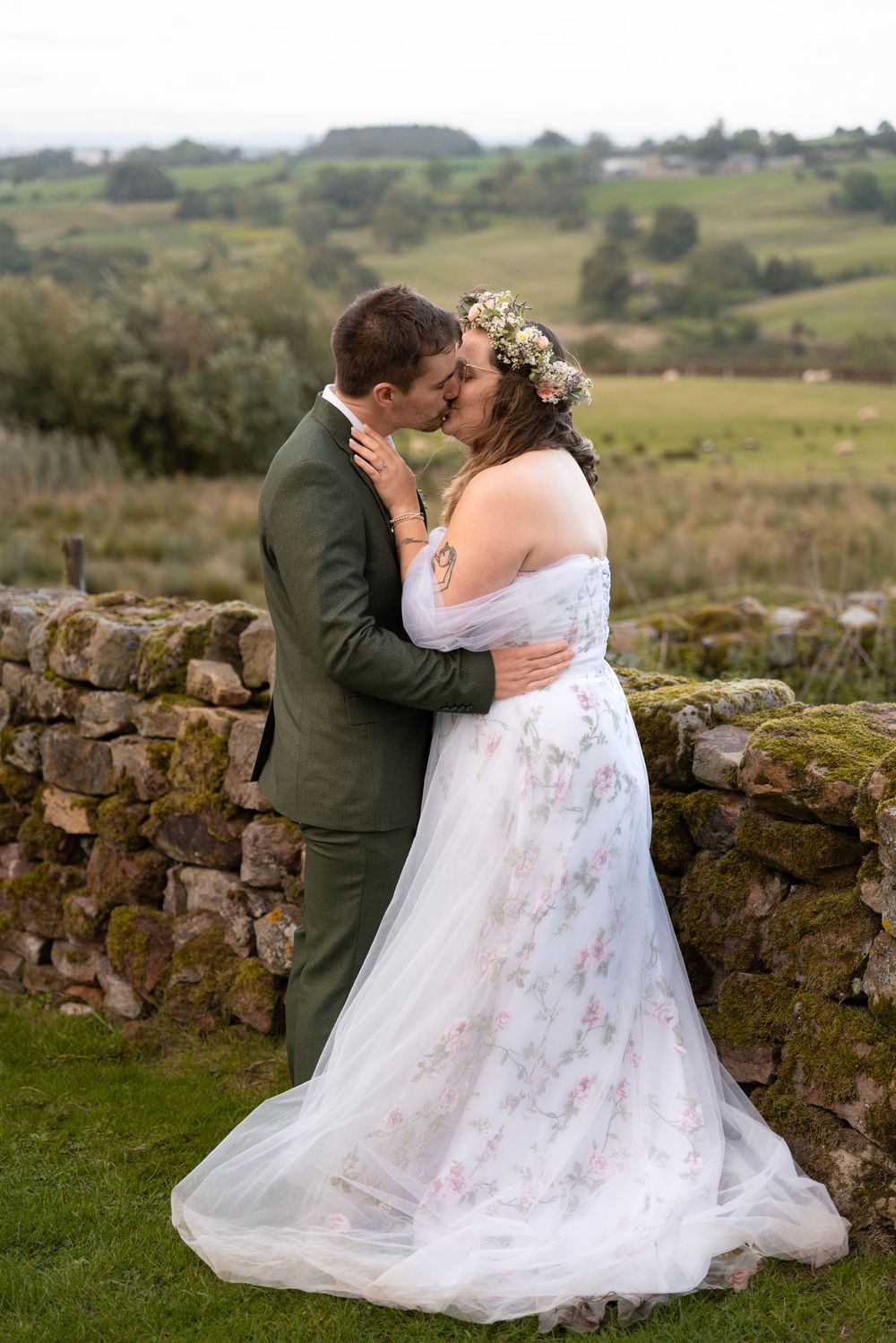 A romantic wedding couple embraces near a rustic stone wall in a scenic countryside setting.