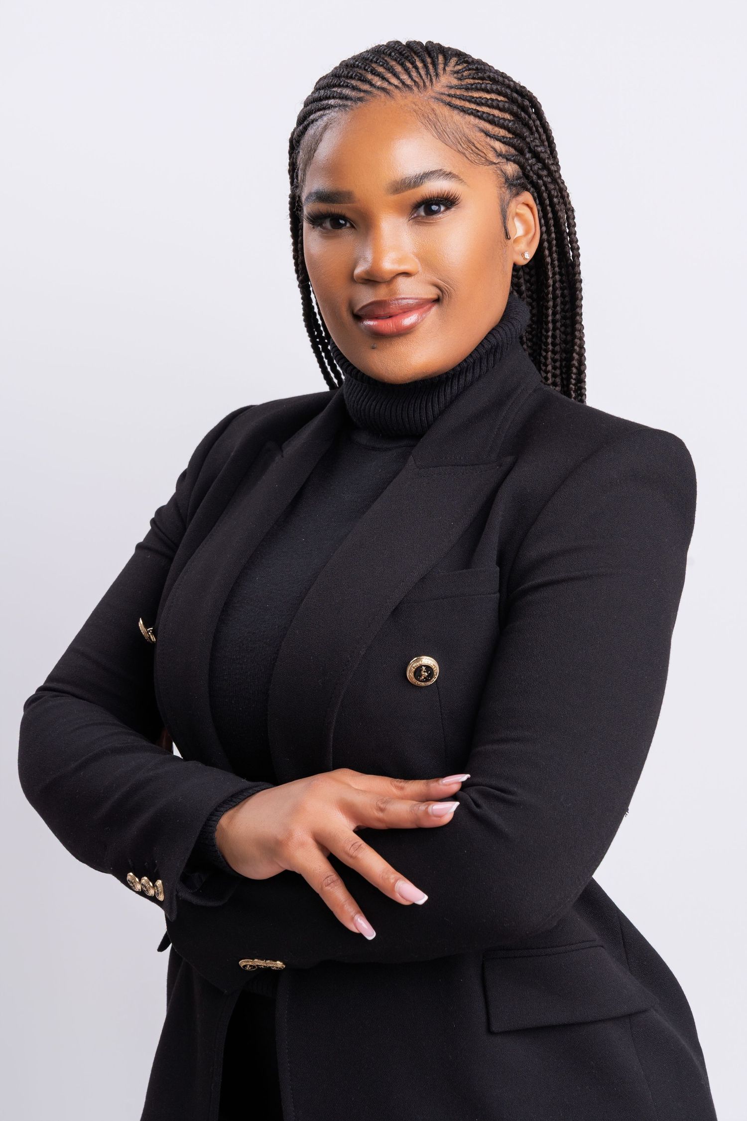 Professional portrait of someone in a black blazer and turtleneck with braided hair against a light gray background.